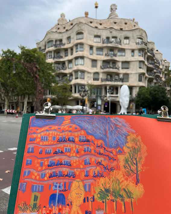 Orange sketchbook page with a drawing of La Pedrera in Barcelona held in front of the building